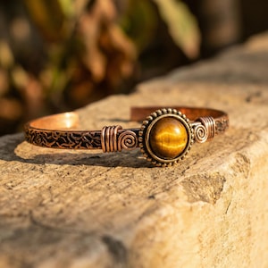 May include: Copper cuff bracelet featuring a large, round tiger's eye stone. The bracelet has intricate wirework and a decorative pattern along the band. The stone is set in a beaded bezel, adding to the bracelet's unique design.