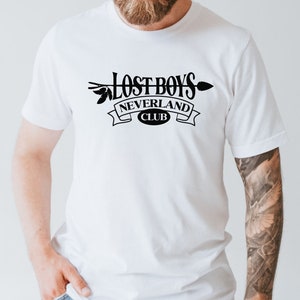 Puede incluir: Camiseta blanca con un dise&ntilde;o gr&aacute;fico negro que dice "Lost Boys Neverland Club" con una flecha y hojas.