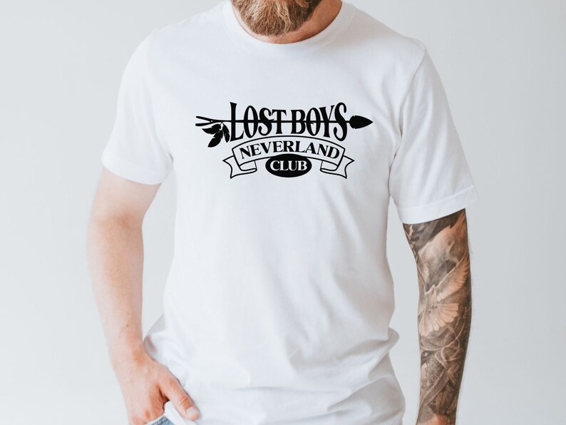 Puede incluir: Camiseta blanca con un dise&ntilde;o gr&aacute;fico negro que dice "Lost Boys Neverland Club" con una flecha y hojas.