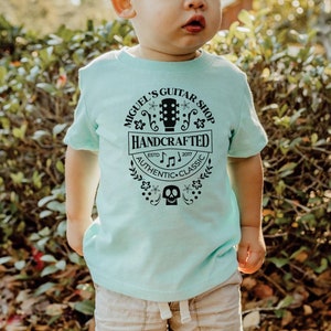 Puede incluir: Una camiseta azul claro con un dise&ntilde;o gr&aacute;fico negro que dice "Miguel's Guitar Shop Handcrafted Est. 2007 Authentic Classic". El gr&aacute;fico tambi&eacute;n incluye una guitarra, notas musicales y una calavera.