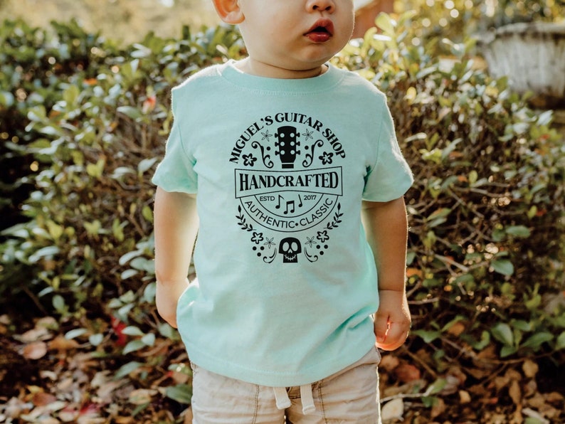 Puede incluir: Una camiseta azul claro con un dise&ntilde;o gr&aacute;fico negro que dice "Miguel's Guitar Shop Handcrafted Est. 2007 Authentic Classic". El gr&aacute;fico tambi&eacute;n incluye una guitarra, notas musicales y una calavera.
