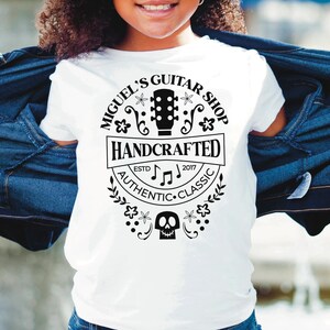 Puede incluir: Una camiseta blanca con un dise&ntilde;o gr&aacute;fico negro que dice "Miguel's Guitar Shop" con una guitarra, flores y las palabras "Handcrafted, Authentic, Classic" y "Est. 2017".