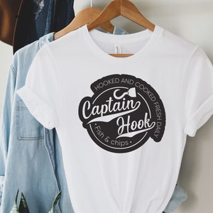 May include: White t-shirt with a black circle graphic that says "Hooked and Cooked Fresh Daily" with a fish hook and the words "Captain Hook" in a stylized font. The text below the circle says "Fish & Chips."