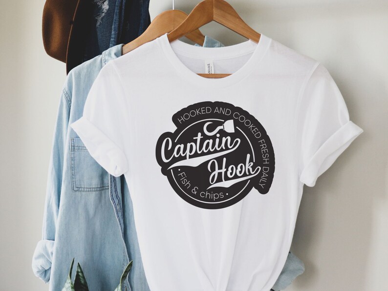 May include: White t-shirt with a black circle graphic that says "Hooked and Cooked Fresh Daily" with a fish hook and the words "Captain Hook" in a stylized font. The text below the circle says "Fish & Chips."