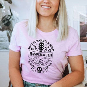 Puede incluir: Una camiseta rosa con un dise&ntilde;o gr&aacute;fico negro que dice "Miguel's Guitar Shop Handcrafted Est. 2017 Authentic Classic" con una guitarra, notas musicales y una calavera.