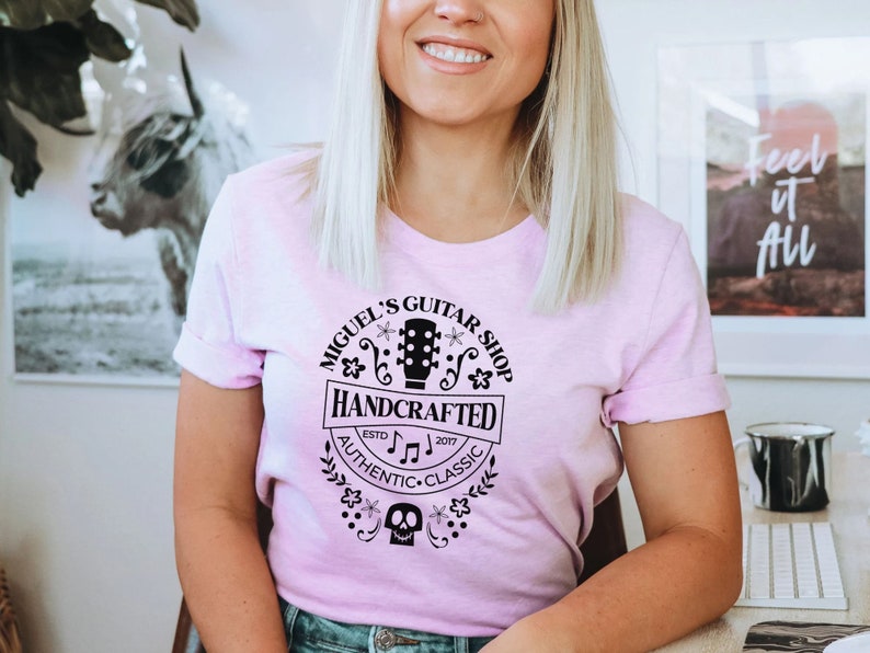 Puede incluir: Una camiseta rosa con un dise&ntilde;o gr&aacute;fico negro que dice "Miguel's Guitar Shop Handcrafted Est. 2017 Authentic Classic" con una guitarra, notas musicales y una calavera.