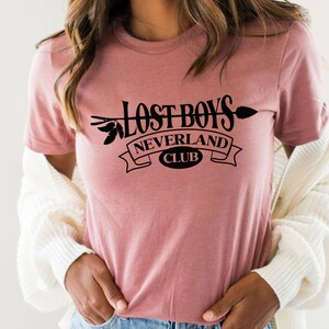 Puede incluir: Una camiseta rosa con un gr&aacute;fico negro que dice "Lost Boys Neverland Club" con una flecha y hojas.