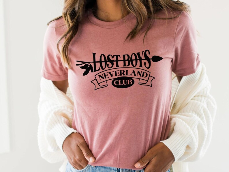 Puede incluir: Una camiseta rosa con un gr&aacute;fico negro que dice "Lost Boys Neverland Club" con una flecha y hojas.