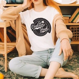 May include: A white t-shirt with a black and white logo that says "Hooked and Cooked Fresh Daily Captain Hook Fish & Chips".