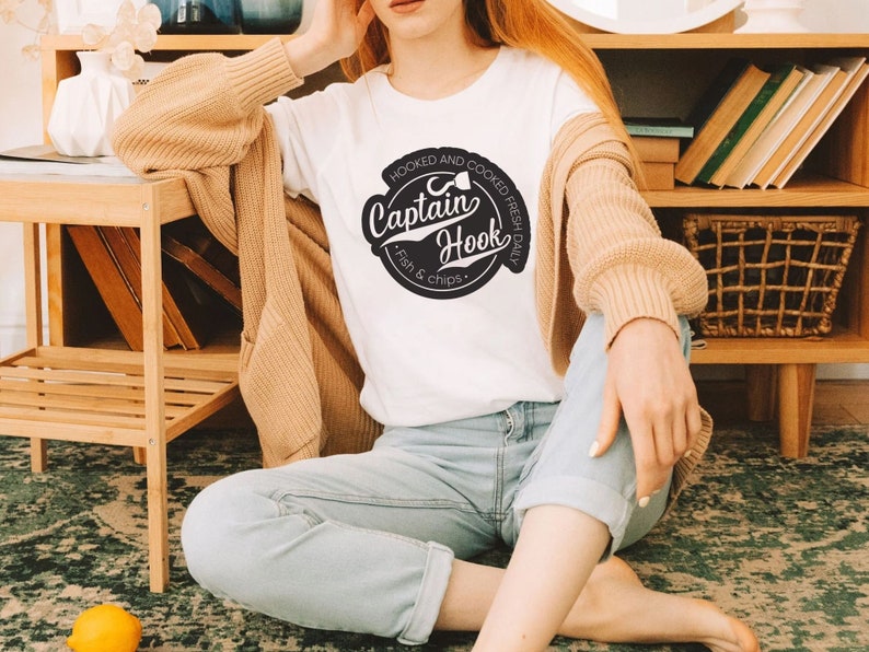 May include: A white t-shirt with a black and white logo that says "Hooked and Cooked Fresh Daily Captain Hook Fish & Chips".