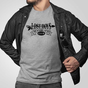 Puede incluir: Una camiseta gris con un gr&aacute;fico negro que dice "Lost Boys Neverland Club" con una flecha y hojas.