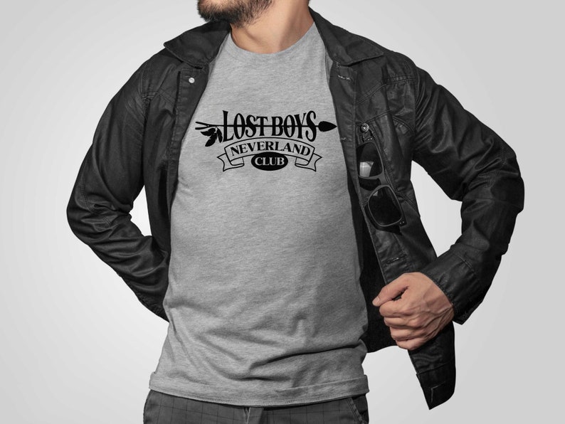 Puede incluir: Una camiseta gris con un gr&aacute;fico negro que dice "Lost Boys Neverland Club" con una flecha y hojas.
