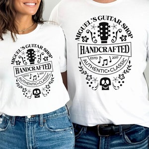 Puede incluir: Dos camisetas blancas con un dise&ntilde;o gr&aacute;fico en blanco y negro que presenta una guitarra, notas musicales, flores y una calavera. El texto en el gr&aacute;fico dice "Miguel's Guitar Shop", "Handcrafted", "Est. 2017", "Authentic" y "Classic".