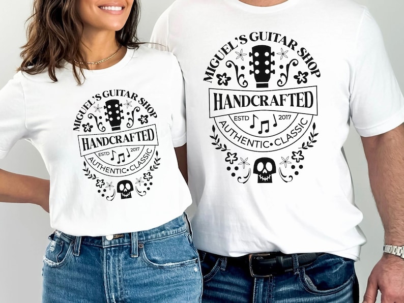 Puede incluir: Dos camisetas blancas con un dise&ntilde;o gr&aacute;fico en blanco y negro que presenta una guitarra, notas musicales, flores y una calavera. El texto en el gr&aacute;fico dice "Miguel's Guitar Shop", "Handcrafted", "Est. 2017", "Authentic" y "Classic".