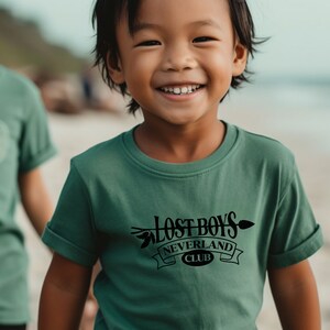 Puede incluir: Un ni&ntilde;o peque&ntilde;o con una camiseta verde con un gr&aacute;fico negro que dice "Lost Boys Neverland Club". El ni&ntilde;o sonr&iacute;e y est&aacute; de pie en una playa.