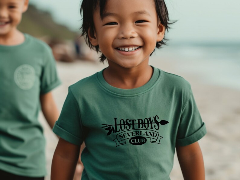 Puede incluir: Un ni&ntilde;o peque&ntilde;o con una camiseta verde con un gr&aacute;fico negro que dice "Lost Boys Neverland Club". El ni&ntilde;o sonr&iacute;e y est&aacute; de pie en una playa.