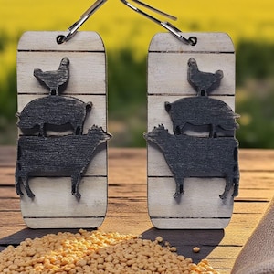 May include: Two wooden earrings with a farm animal theme. Each earring features a black silhouette of a cow, pig, and chicken stacked on top of each other. The background is a blurred image of a field and a red barn.