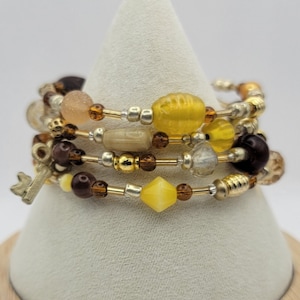 Könnte beinhalten: Ein Set aus vier Armbändern mit braunen, goldenen und gelben Perlen. Die Armbänder sind auf einem weißen Präsentationsständer mit Holzsockel gestapelt. Der Sockel trägt ein Etikett mit der Aufschrift "F-5".