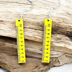 以下が含まれることがあります： 黄色の定規のデザインが特徴的な、ペアの個性的なイヤリング。各イヤリングにはシルバーのフックと、1から10までの黒いマークと数字が付いた長方形の黄色のペンダントが付いています。イヤリングは流木の上に展示されています。