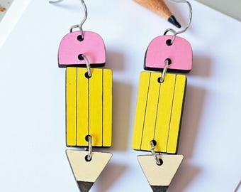 May include: A pair of whimsical earrings shaped like pencils. Each earring features a pink eraser top, a yellow pencil body with black lines, and a cream-colored tip with a black point. The earrings hang from silver-colored hooks.