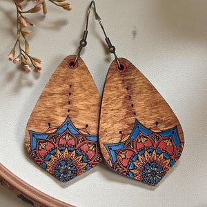 Peut inclure: Une paire de boucles d'oreilles en bois en forme de goutte d'eau, ornées d'un motif mandala détaillé dans des tons de bleu, rouge et jaune. Les boucles d'oreilles présentent une finition en grain de bois naturel et des crochets en métal sombre.