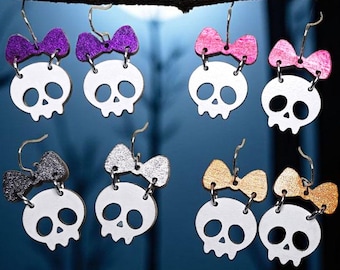 May include: A collection of Halloween-themed earrings. Each earring features a white skull with a decorative bow in various colors, including purple, pink, silver, and gold. The earrings are suspended from a branch against a dark, moonlit background.