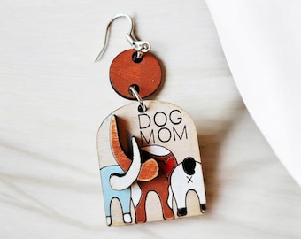 Puede incluir: Pendientes de madera con un círculo marrón, un arco marrón claro con el texto "DOG MOM" y tres perros de dibujos animados en azul, marrón y blanco.