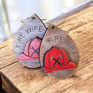 May include: Two wooden teardrop-shaped earrings. Each earring features a firefighter's helmet design with the words "FIRE WIFE" engraved. One helmet is red, the other is pink, both with crossed axes. The earrings are on a wooden surface.