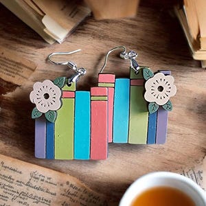 Puede incluir: Pendientes con forma de libros coloridos con un adorno floral. Los pendientes presentan rayas verticales en rosa, azul, verde y morado. El adorno floral es de color beige claro con hojas verdes. Sobre una superficie de madera.