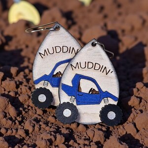 May include: Wooden teardrop-shaped earrings with a blue truck design and the word "Muddin'" in black. The truck has large black tires with silver centers. The earrings are on a brown surface.