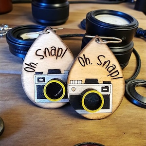 May include: Two wooden teardrop-shaped keychains with a camera design. Each keychain has the words "Oh. Snap!" etched into the wood. The camera design features a black body with a yellow lens.