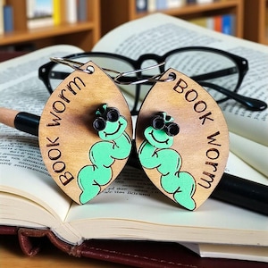 May include: Wooden leaf-shaped earrings with the words "BOOK WORM" engraved on them. Each earring features a green cartoon worm with black glasses. The earrings are displayed on an open book with glasses and a pen in the background.