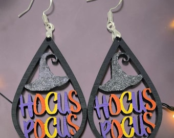 Könnte beinhalten: Ein Paar tropfenförmige Ohrringe mit einem schwarzen Rahmen und einem glitzernden grauen Hexenhut an der Oberseite. Der Schriftzug HOCUS POCUS ist in der Mitte ausgeschnitten, wobei die Buchstaben abwechselnd in Lila, Orange und Gelb gehalten sind.