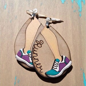 May include: Wooden teardrop earrings with a runner design. The earrings show a leg with a running shoe in teal, purple, and white. The word "Runner" is written in cursive on the earrings. The background is a textured tan surface with teal accents.