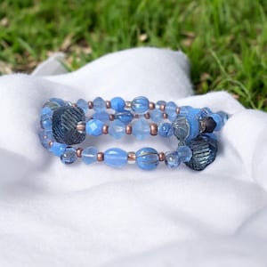 Könnte beinhalten: Ein dreireihiges Armband aus blauen und kupferfarbenen Glasperlen. Die Perlen sind in verschiedenen Formen und Größen, darunter rund, oval und facettiert.