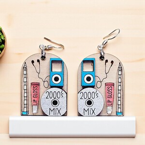 May include: Wooden earrings featuring a blue MP3 player, a CD with "2000's MIX" text, and a tube of lip gloss. The earrings also include a drawing of headphones and a pencil. The earrings are on a white surface.