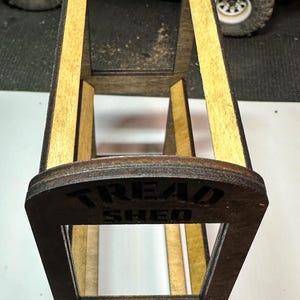 Op de afbeelding: Een houten displaystandaard met de tekst "TREAD SHED" op de voorkant. De standaard heeft een donkerbruin frame met lichtbruine binnensteunen. Het ontwerp is een miniatuurreplica van een grotere structuur, waarschijnlijk voor modelauto's of vergelijkbare items.