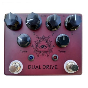 May include: A red guitar pedal with the text "DUAL DRIVE" and a black and white illustration of an eye inside a triangle with flames. The pedal has four knobs labeled "D", "V", "Tone", and "Tone", and two footswitches.