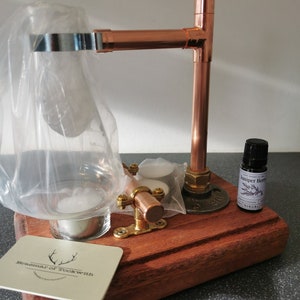 May include: A copper and wood essential oil diffuser with a glass jar, a metal ring, and a small bottle of juniper berry essential oil. The diffuser is on a wooden base with a business card that says "Braemar of Tolkwith".