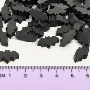 May include: A collection of small, black bat-shaped embellishments scattered on a white surface. A purple ruler with centimeter markings is in the foreground. These decorative pieces are ideal for crafting and Halloween-themed projects.
