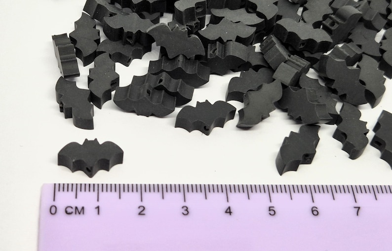 May include: A collection of small, black bat-shaped embellishments scattered on a white surface. A purple ruler with centimeter markings is in the foreground. These decorative pieces are ideal for crafting and Halloween-themed projects.