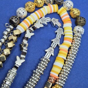 May include: An assortment of beaded jewelry components on a blue surface. The components include silver, gold, and black beads, along with bracelets featuring butterfly and Hamsa hand charms. There are also colorful polymer clay disc beads.