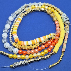 May include: A collection of colorful beaded necklaces and bracelets. The jewelry features a variety of beads in shades of yellow, orange, white, silver, and gold. Some beads are geometric, while others are round or disc-shaped. The background is a solid blue.