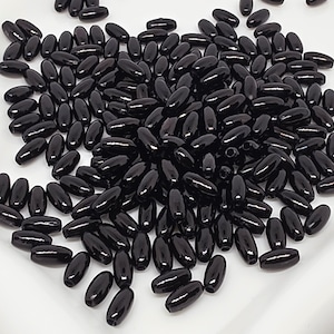 May include: A large collection of small, smooth, glossy black beads with an elongated oval shape. Each bead features a central hole running through its length, and they are scattered in a loose pile, showcasing their uniform dark colour and reflective surface.