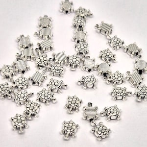 May include: A collection of small, silver-coloured turtle charms. The charms have detailed shell patterns and are scattered across a white surface. These decorative charms are suitable for jewellery making or craft projects.
