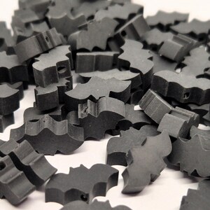 May include: A pile of small, dark gray bat-shaped wooden cutouts. Each cutout has a detailed wing design and a small hole. The bats are scattered on a white surface, creating a textured, Halloween-themed display.