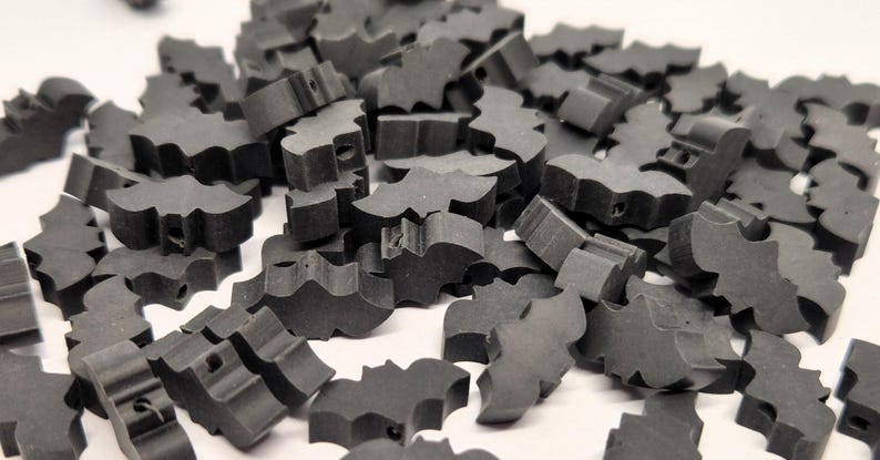 May include: A pile of small, dark gray bat-shaped wooden cutouts. Each cutout has a detailed wing design and a small hole. The bats are scattered on a white surface, creating a textured, Halloween-themed display.
