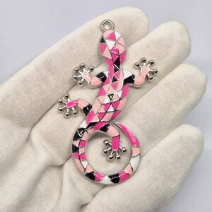 May include: A vibrant lizard pendant with a geometric pattern. The pendant showcases a pink, black, and white design on its body and a silver-coloured loop for hanging. The lizard has detailed feet and a curled tail.