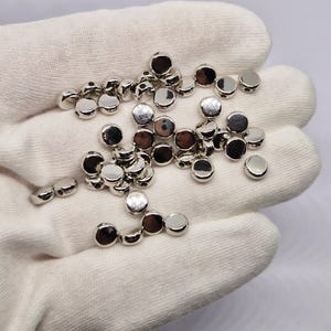 May include: A collection of small, shiny, silver-coloured metal beads. The beads are round and flat, with a small hole on each side. They are scattered on a white fabric surface.