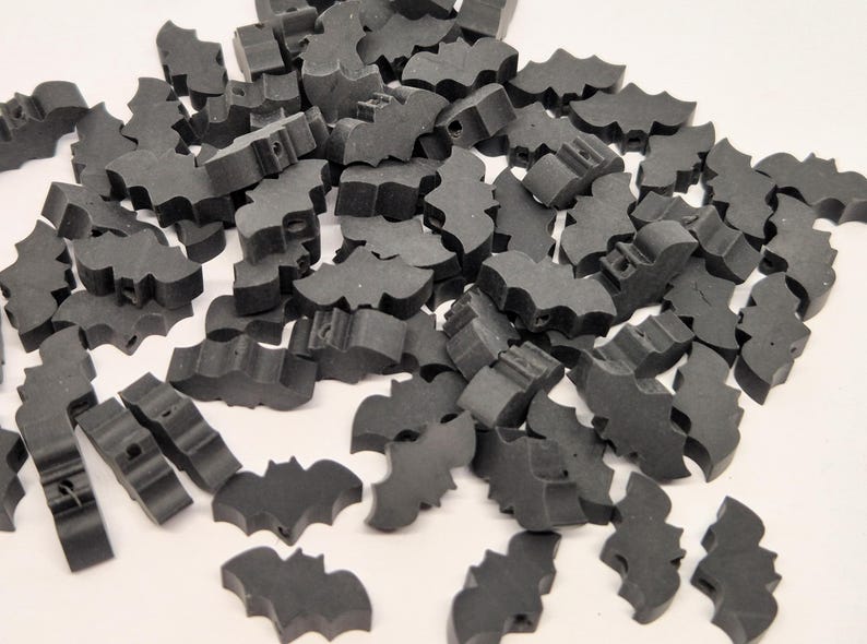 May include: A pile of small, dark gray bat-shaped wooden beads. Each bead has a hole for stringing and is designed with a bat silhouette. These beads are suitable for crafting and jewelry making.